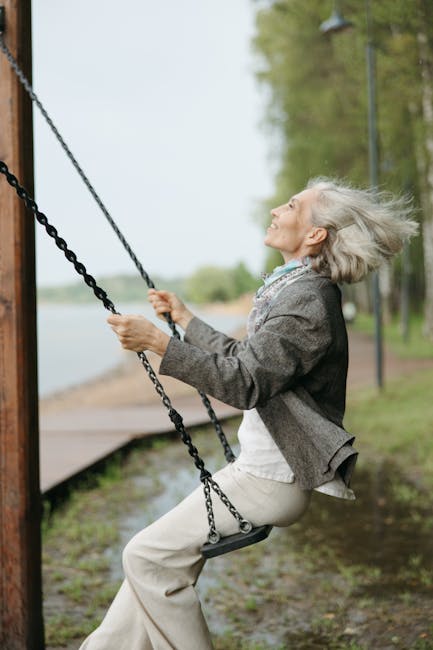 A joyful senior woman swinging in a park, surrounded by nature on a bright day.
