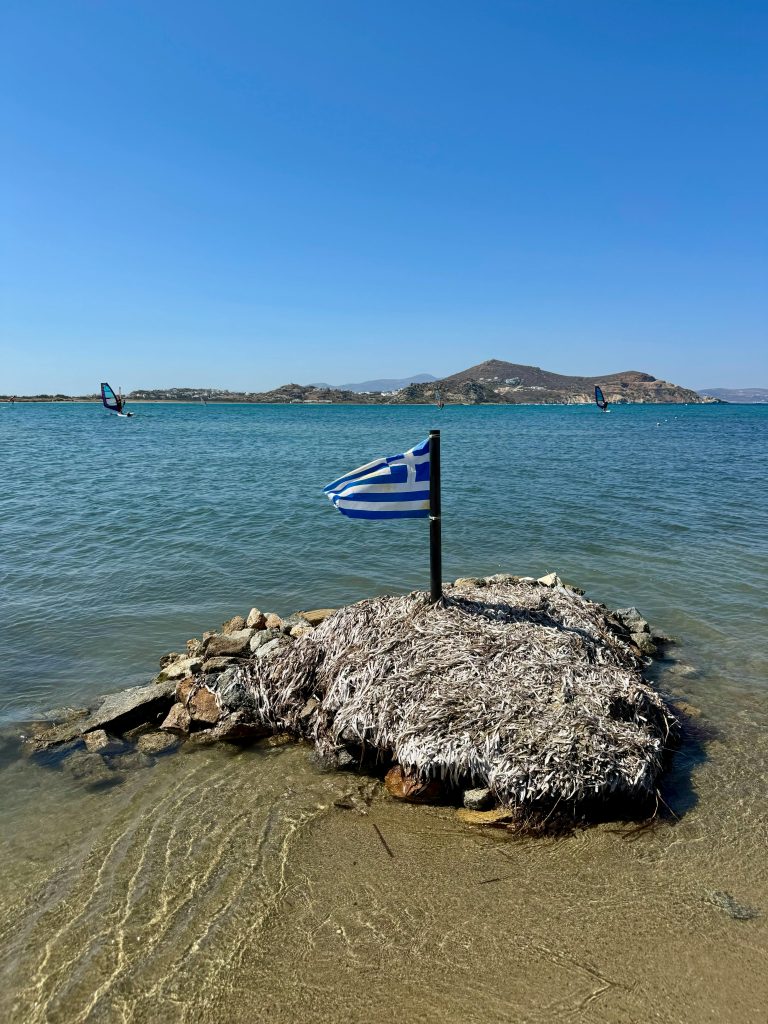 A Greek flag stands on a small beach island with a picturesque sea backdrop.