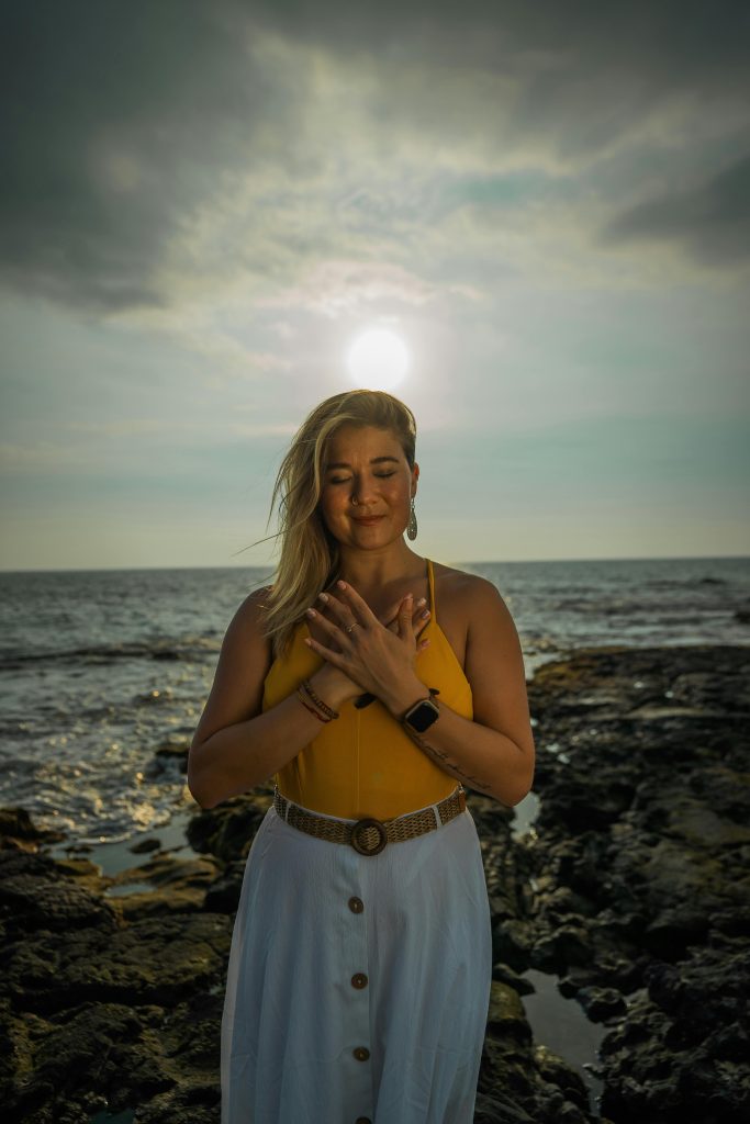 A woman practices mindfulness and meditation by the ocean during sunset, embodying peace and wellness.