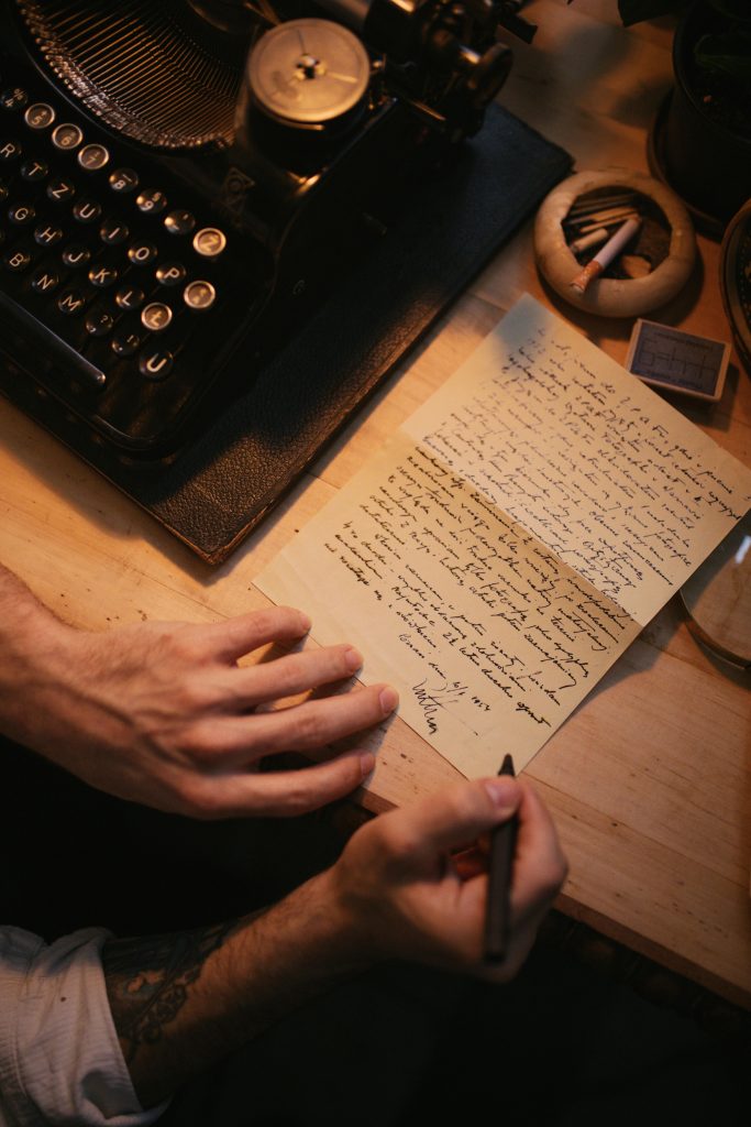 Close-up of hands writing a letter next to a vintage typewriter on a wooden desk.