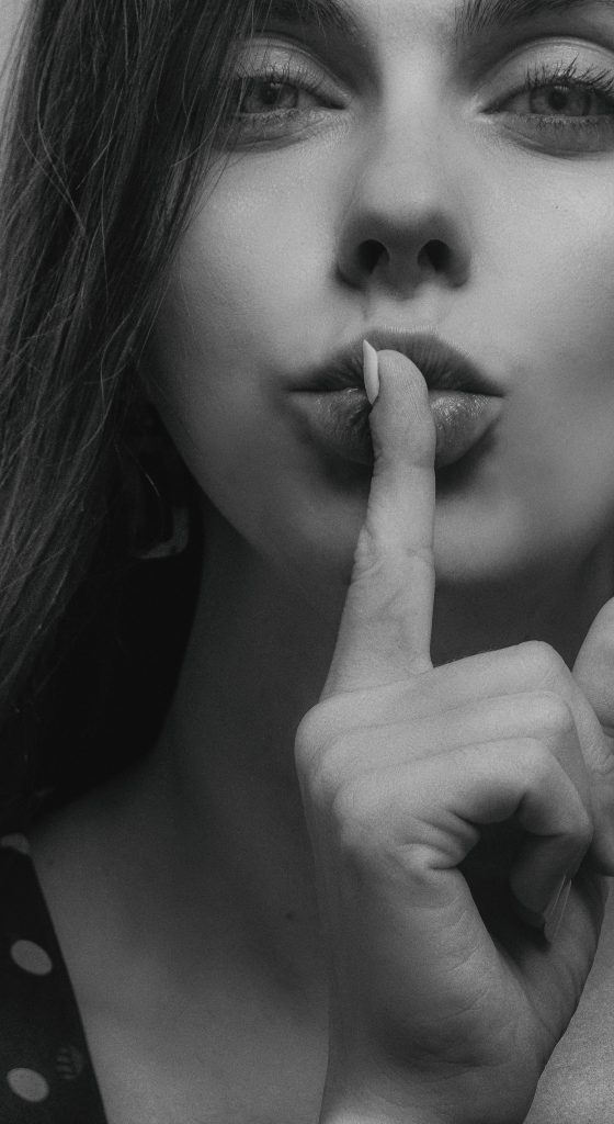 Close-up black and white portrait of a woman gesturing silence with a finger over lips.