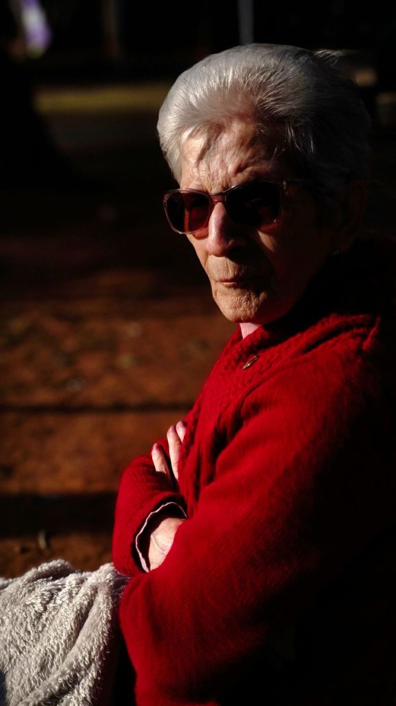 Senior woman enjoying a sunny day in the park, dressed warmly and wearing sunglasses.